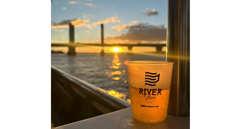 Copo do River Beer no parapeito do catamarã com o sol se pondo atrás da ponte Presidente Dutra